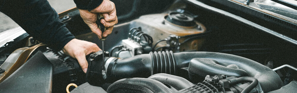 Ein Mechaniker repariert den Motor eines Autos, indem er mit Werkzeug einige Teile anpasst. Im Bild ist ein offener Motorraum mit verschiedenen Autoteilen zu sehen.